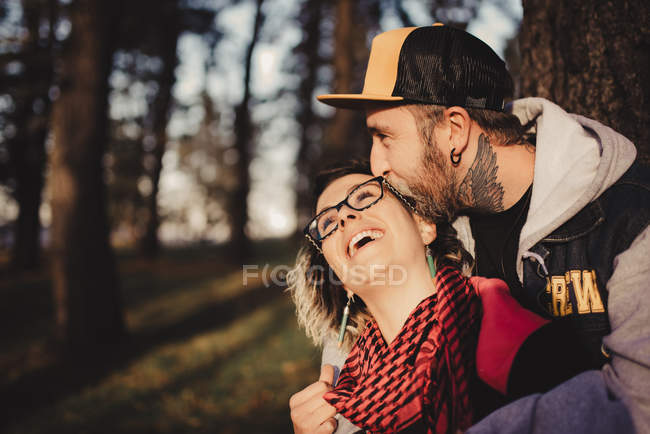 Uomo barbuto abbracciando dalla schiena donna allegra vicino legno nella foresta su sfondo sfocato — Foto stock