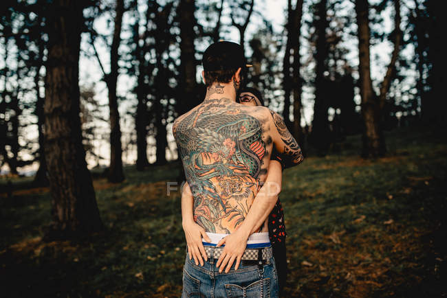 Vista posterior de un hombre joven sin camisa en tatuajes con una mujer abrazada en el parque sobre un fondo borroso - foto de stock