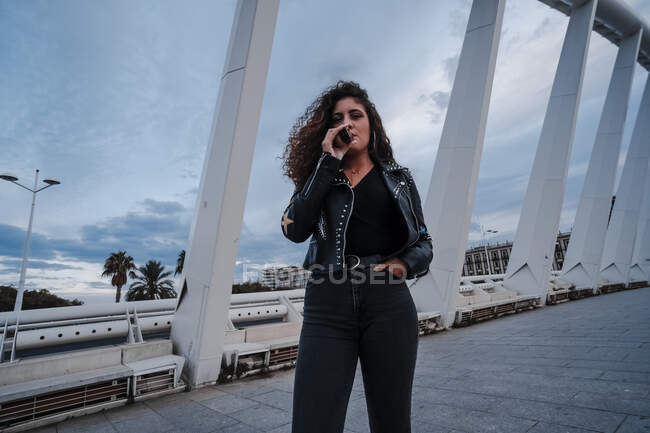 Стильная женщина в черной куртке и джинсах курит на мосту — стоковое фото
