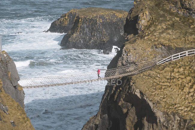 Tourist walking on rope bridge suspended between cliffs in Northern Ireland — Stock Photo