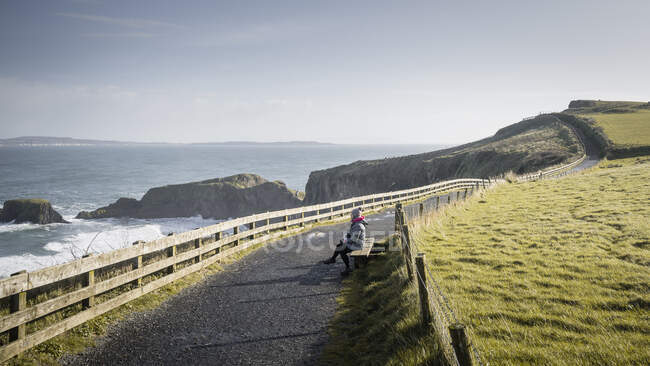 Жінка сидить на лавці на сільській дорозі між морем і полем — стокове фото