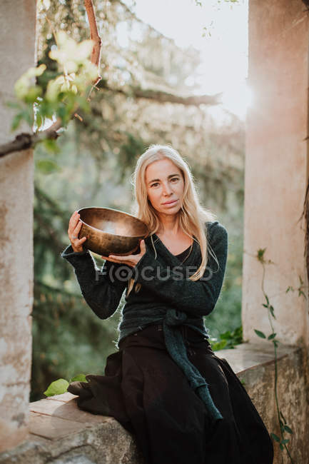Woman in long gown clothes looking at camera and holding metal musical bowl while sitting in ruins of old castle in historic place — Stock Photo