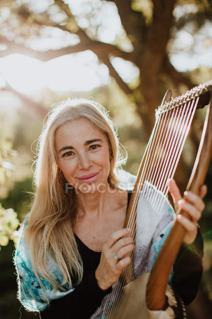 Романтическая очаровательная женщина с светлыми волосами, наслаждающаяся мелодией, играя на музыкальном инструменте и сидя в саду летом — стоковое фото