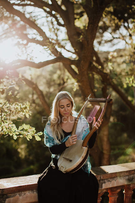 Romantique charmante femme aux cheveux blonds jouissant de la mélodie tout en jouant de l'instrument de musique et assis dans le jardin en été — Photo de stock