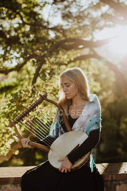 Romantique charmante femme aux cheveux blonds jouissant de la mélodie tout en jouant de l'instrument de musique et assis dans le jardin en été — Photo de stock