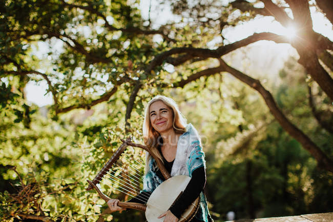 Musician wearing scarf on shoulders while holding lyre and standing green garden in summer day in back lit — Stock Photo