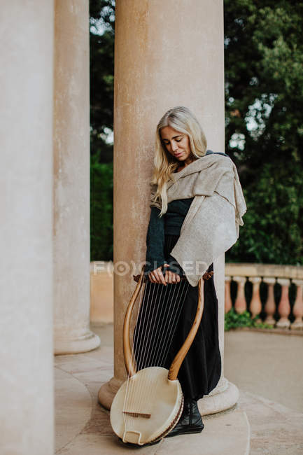 Female musician in casual clothes resting after performance and holding wooden lyre while leaning on pillar at building entrance — Stock Photo
