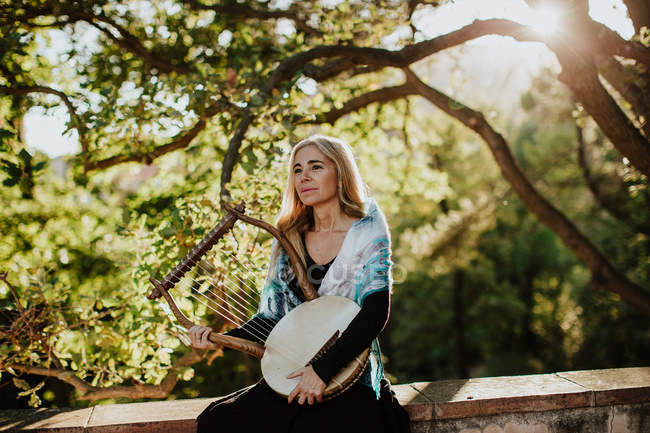 Жінка-музикантка в шарфі на плечах, тримаючи ліру і стоячи зелений сад в літній день в спині освітлений — стокове фото