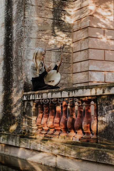 Seitenansicht einer Frau, die an einem Musikinstrument spielt, während sie an einem sonnigen Tag auf dem Geländer eines alten Steingebäudes sitzt — Stockfoto