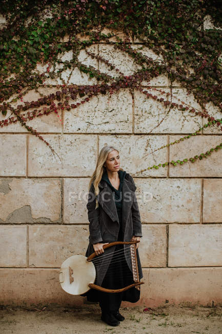 Thoughtful charming blond female musician in coat holding string musical instrument while standing at old stone wall in autumn — Stock Photo