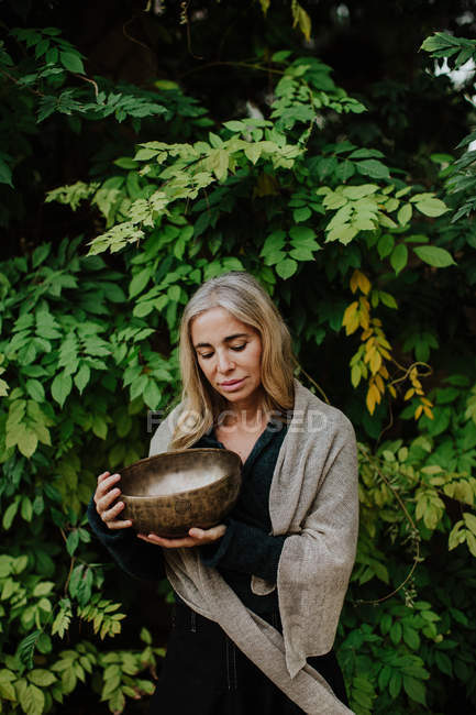 Woman in long gown clothes with closed eyes and holding metal musical bowl while sitting in ruins of old castle in historic place — Stock Photo