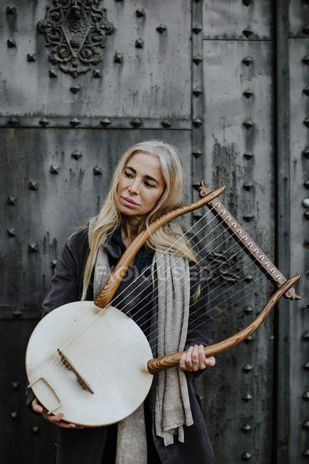 Задумчивая привлекательная блондинка-музыкантка нежно держит струнный музыкальный инструмент, стоя у металлических ворот — стоковое фото
