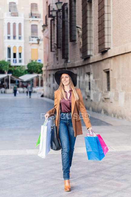 Femme satisfaite en tenue décontractée élégante et chapeau marchant avec des sacs à sauter et regardant loin de la rue de la ville — Photo de stock
