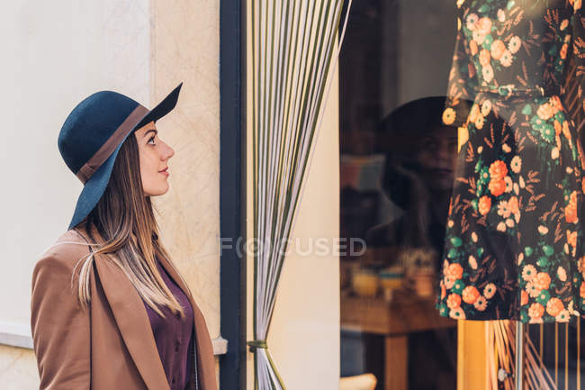 Side view of young woman in stylish hat and brown jacket looking through shop window on fashionable dress with roses — Stock Photo