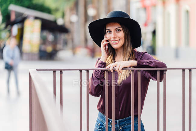 Zufriedene langhaarige Frau mit modischem schwarzen Hut und Hemd lehnt am Zaun, während sie mit dem Handy telefoniert und in die Kamera schaut — Stockfoto