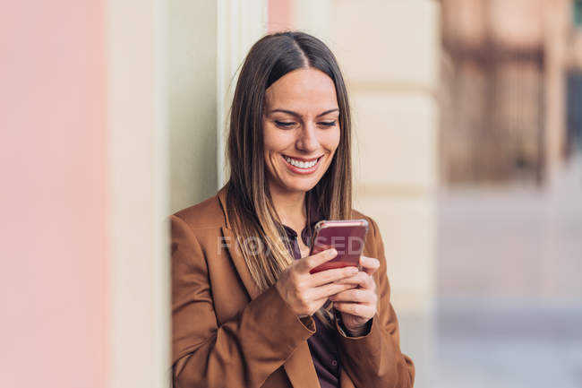 Satisfied charming woman in stylish casual clothing and heels leaning on wall of building using mobile phone — Stock Photo