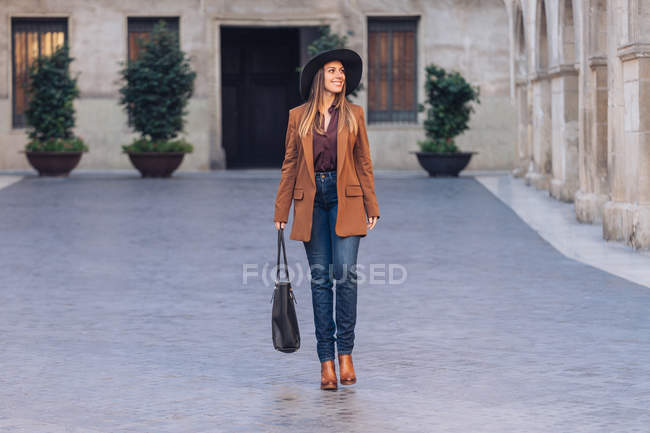 Возбужденная женщина в стильной повседневной одежде и черной шляпе, идущая и смотрящая на тротуар среди старых зданий — стоковое фото