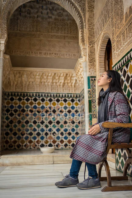 Vista lateral de la elegante mujer asiática sentada en la silla y descansada dentro del antiguo palacio islámico. - foto de stock