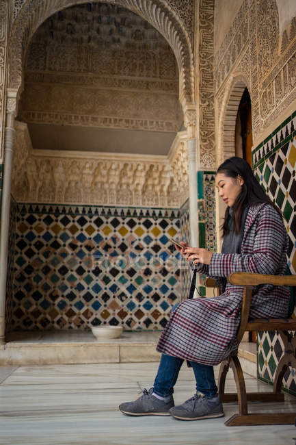 Вид сбоку на стильную женщину, сидящую на стуле и отдыхающую внутри старого исламского дворца с помощью мобильного телефона — стоковое фото
