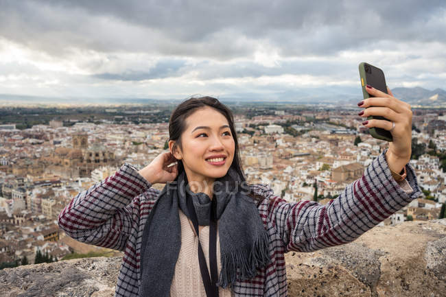 Feliz mujer con un estiloso equipo que toca el cabello y se toma selfie mientras se enfrenta a la ciudad vieja y el cielo nublado. - foto de stock