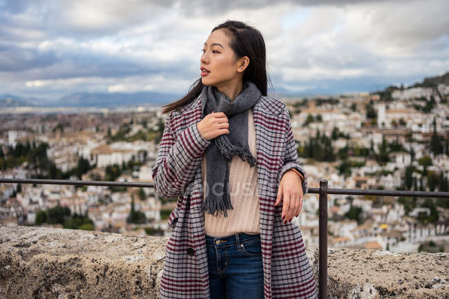 Щаслива азіатська жінка в стильному пальто і шарф посміхається і дивиться геть, стоячи на розмитому тлі старого міста — стокове фото
