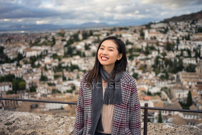 Счастливая женщина в стильном пальто и шарфе улыбается и смотрит в камеру, стоя на размытом фоне старого города — стоковое фото