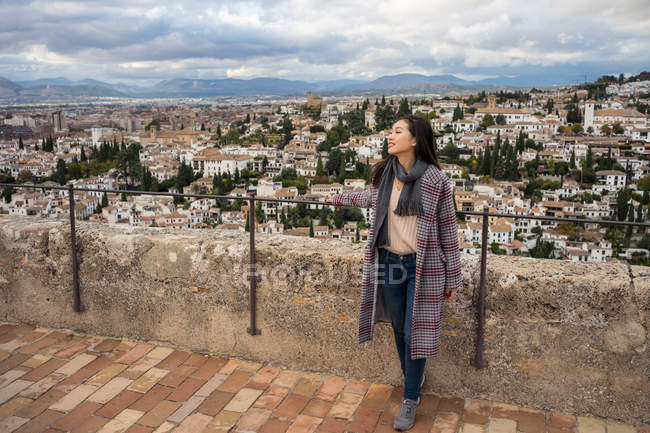 Glückliche asiatische Frau in stylischem Mantel und Schal lächelt und schaut weg, während sie auf verschwommenem Hintergrund der gealterten Stadt steht — Stockfoto