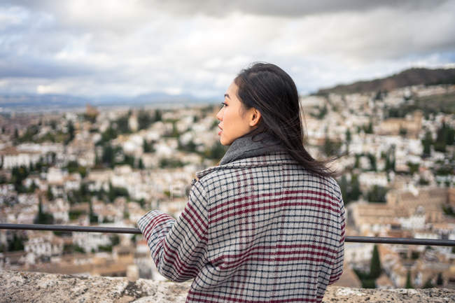 Вид сзади на женщину в стильном пальто и шарфе, улыбающуюся и отводящую взгляд, стоя на размытом фоне старого города — стоковое фото