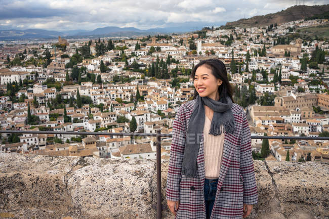 Happy asian woman in stylish coat and scarf smiling and looking away while standing on blurred background of aged city — Stock Photo