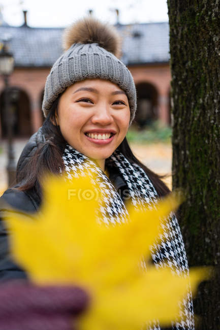 Donna asiatica in abiti caldi guardando macchina fotografica e coprendo faccia con foglia di acero giallo mentre in piedi alla Cattedrale di Oslo in Norvegia — Foto stock