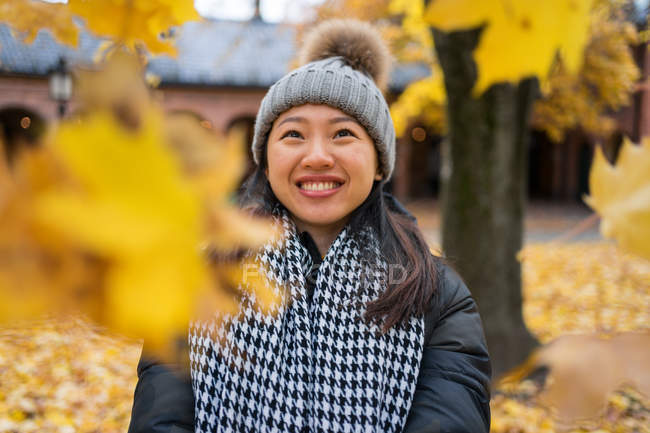 Jovem asiática alegre jogando folhas amarelas de bordo enquanto está em pé no pátio da Catedral de Oslo, na Noruega olhando para longe — Fotografia de Stock