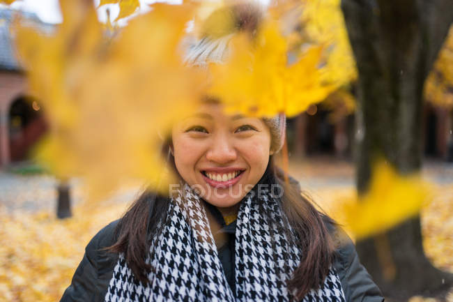 Una mujer asiática alegre tirando hojas de arce amarillo mientras se encuentra en el patio de la catedral de Oslo, en Noruega, mirando hacia otro lado. - foto de stock