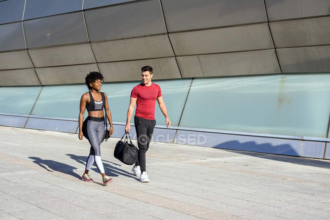 Donna afroamericana e uomo caucasico con borsa sportiva in mano camminando insieme lungo la parete della costruzione della città — Foto stock