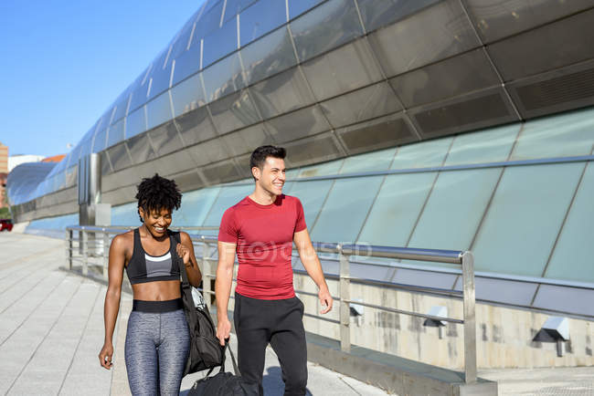 African American woman and caucasian man with sports bag in hand walking together along wall of city construction — Stock Photo