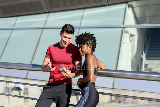 Personal Trainer mit Smartphone und zeigt einer lächelnden Afroamerikanerin App, während sie neben dem Gebäude zusammen steht — Stockfoto