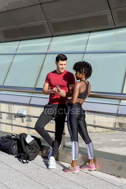 Entraîneur personnel utilisant un smartphone et montrant une femme afro-américaine souriante tout en se tenant debout à côté du bâtiment — Photo de stock