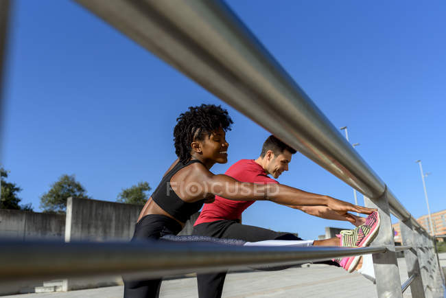 Vista a basso angolo della donna afroamericana e sorridente uomo sportivo in piedi accanto alla recinzione metallica e gambe stiranti — Foto stock