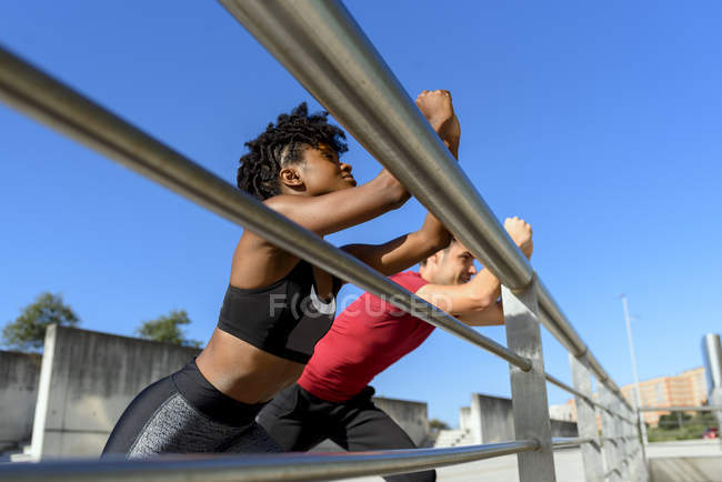 Low angle view of African American woman and smiling sportive man standing next to metal fence and stretching legs — Stock Photo