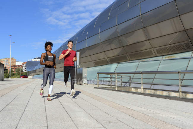 Athletische Afroamerikanerin in Top und Leggings joggt mit sportlichem männlichen Freund neben modernem Stadtbau — Stockfoto