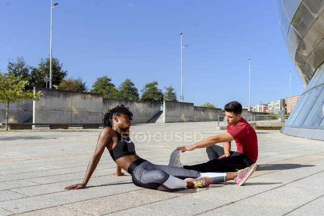 African American woman and caucasian man sitting and stretching forward while practicing together in city in summer day — Stock Photo