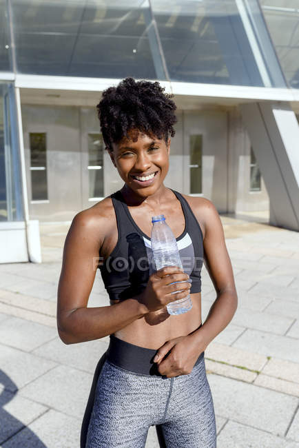 African American woman in sports top standing next to building and holding bottle of water while having break during training — Stock Photo