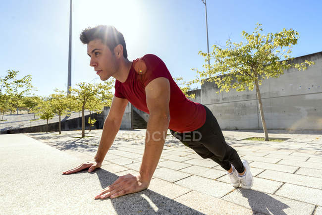 Vista lateral do homem vestindo camisa vermelha e calças pretas fazendo push up ou exercício de prancha ao lado de banco de concreto na cidade na parte de trás iluminado — Fotografia de Stock
