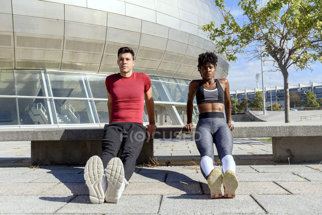 African American sportswoman and caucasian sportsman in red shirt doing push up exercises next to concrete bench in city — Stock Photo