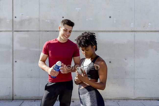 Веселый мужчина с бутылкой воды и афроамериканка делят смартфон стоя — стоковое фото