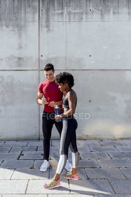 Веселий чоловік з пляшкою води і афро-американська жінка поділяють смартфон стоячи разом. — стокове фото