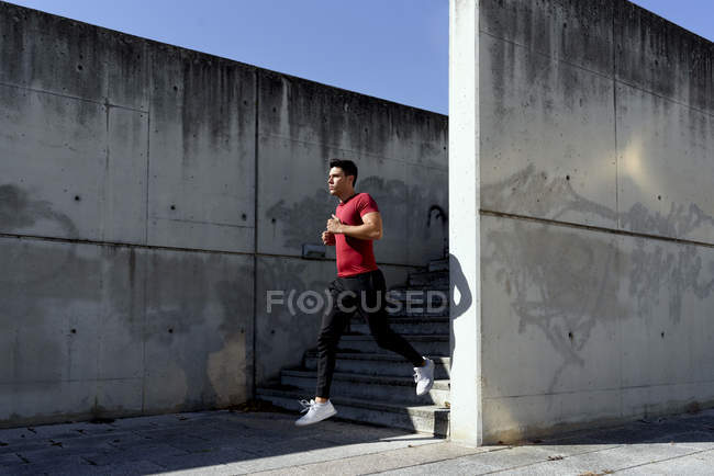 Hombre con camisa roja y pantalones negros bajando las escaleras durante el entrenamiento en el soleado día de verano en la ciudad. - foto de stock