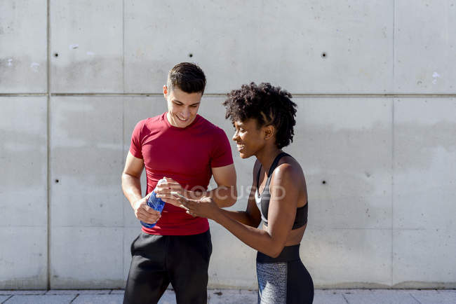 Allegro caucasico uomo con bottiglia d'acqua e afroamericana donna condivisione smartphone in piedi insieme — Foto stock