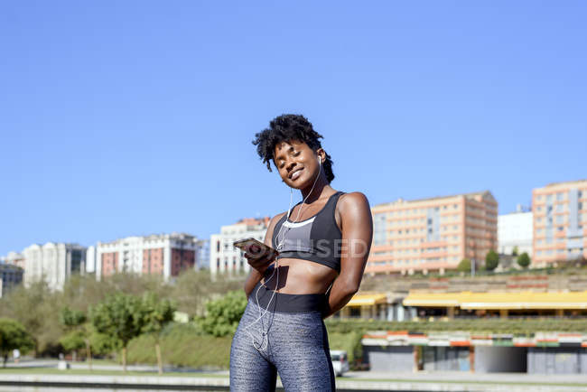 Mujer afroamericana en el deporte que escucha música con teléfonos inteligentes y auriculares mientras entrena en la orilla del río en la ciudad. - foto de stock