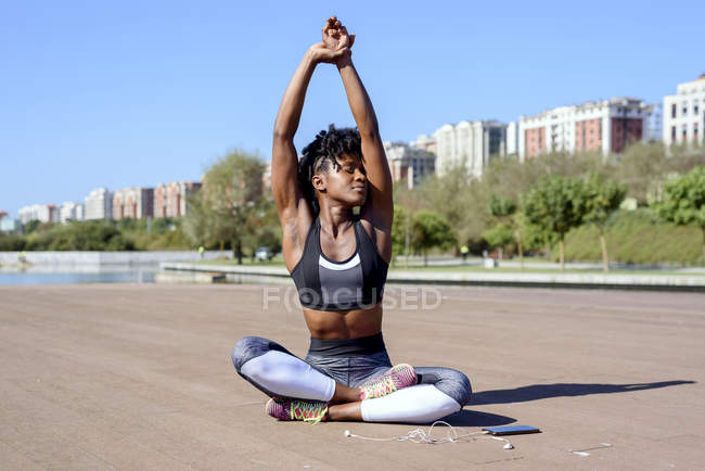 Афроамериканська жінка на спортивному вершку і лаки сидять у позі лотоса і розтягують руки вгору під час відпочинку. — стокове фото