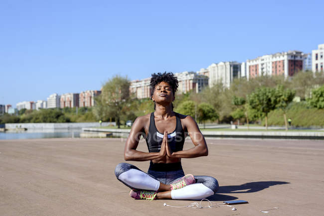 Sportive afro-américaine moderne assise dans un lotus posant et méditant après un entraînement au bord de la rivière en ville en été — Photo de stock